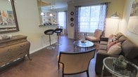 This single-floor space showcases an open-concept layout that seamlessly connects a bar area to a comfortable living space. The rich, dark hardwood flooring runs throughout, providing a warm and cohesive foundation for the interior. A built-in bar counter with sleek, adjustable black bar stools and overhead pendant lighting creates a social focal point, complemented by open shelving displaying decorative accents. The living area is bathed in natural light courtesy of large windows dressed with patterned grommet-top curtains in a neutral palette, offering views of the surrounding greenery. Elegant upholstered accent chairs in a warm beige tone are arranged around a glass-top coffee table, creating an inviting conversational setting. Wall-mounted decorative plates add a curated, intentional touch to the décor. The space balances functionality and style, with clean lines, modern fixtures, and a cohesive neutral color scheme punctuated by subtle decorative details. The open flow between the bar and living zones enhances the sense of spaciousness, making the layout feel both practical and welcoming.