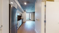 This single-floor space showcases an industrial-modern aesthetic defined by its most striking architectural feature: exposed concrete ceilings that span throughout, lending a raw, loft-like character to the interior. Light hardwood flooring runs continuously across the living and sleeping areas, complementing the clean white walls and creating a warm contrast against the industrial overhead finish. Structural concrete columns are visible within the living area, reinforcing the building's bold architectural identity.

The kitchen is well-appointed with a full suite of stainless steel appliances, including a range with oven, built-in microwave, dishwasher, and refrigerator. Sleek white cabinetry extends to the ceiling, providing generous storage, while dark countertops and a glossy black tile backsplash add a contemporary edge to the space.

Large windows in both the living area and bedroom admit substantial natural light and frame views of the surrounding urban skyline. The bedroom features its own closet with built-in shelving. The bathroom includes a full soaking tub and shower combination with white tile surround and dark tile flooring. Recessed lighting throughout ensures a well-lit environment across all areas of the space.