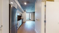 This single-floor space showcases an industrial-modern aesthetic defined by its most striking architectural feature: exposed concrete ceilings that span throughout, lending a raw, loft-like character to the interior. Light hardwood flooring runs continuously across the living and sleeping areas, complementing the clean white walls and creating a warm contrast against the industrial overhead finish. Structural concrete columns are visible within the living area, reinforcing the building's bold architectural identity.

The kitchen is well-appointed with a full suite of stainless steel appliances, including a range with oven, built-in microwave, dishwasher, and refrigerator. Sleek white cabinetry extends to the ceiling, providing generous storage, while dark countertops and a glossy black tile backsplash add a contemporary edge to the space.

Large windows in both the living area and bedroom admit substantial natural light and frame views of the surrounding urban skyline. The bedroom features its own closet with built-in shelving. The bathroom includes a full soaking tub and shower combination with white tile surround and dark tile flooring. Recessed lighting throughout ensures a well-lit environment across all areas of the space.