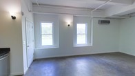 This single-floor space showcases a clean industrial-modern design that balances raw architectural elements with a bright, welcoming atmosphere. The polished concrete floors serve as a defining feature, lending durability and a sleek, contemporary character to the interior. Painted in a soft, neutral gray-white tone, the walls contribute to an open and airy feel throughout the space.

Two double-hung windows are positioned along the main wall, flooding the interior with natural light and offering views of the surrounding greenery outdoors. A wall-mounted light fixture between the windows provides additional illumination, complementing the natural light source. Exposed white-painted pipes run along the ceiling, reinforcing the space's industrial aesthetic while keeping the overall look tidy and intentional.

A wall-mounted split HVAC unit ensures climate control, providing year-round comfort. The ceiling features visible structural beams, adding to the loft-like character of the space. A paneled door on the adjacent wall provides access to additional areas. The combination of raw materials, neutral tones, and natural light creates a versatile and functional environment well-suited for a variety of uses.