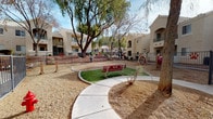 This outdoor dog park is a thoughtfully designed amenity situated within a multi-building residential community featuring Southwest-style stucco architecture with tiled rooflines, arched detailing, and private balconies. The park is fully enclosed by sturdy metal fencing, providing a safe and secure environment for pets to roam and play off-leash.

The grounds are landscaped with a combination of artificial turf patches and natural gravel terrain, offering varied surfaces for activity. A winding concrete pathway meanders through the space, creating a pleasant route for owners to walk alongside their pets. Several mature shade trees are distributed throughout the park, providing welcome canopy cover and a natural focal point around which the pathways curve.

Dog agility equipment is a standout feature of the space, including a jump hoop in a bold red and tan color scheme, as well as additional interactive obstacles. Distinctive red metal benches, each adorned with decorative paw print cutouts, are placed throughout the park, offering comfortable seating for pet owners. Raised planter beds with colorful flowers add a welcoming visual accent near the building entrances. The open-air layout ensures ample natural light throughout the day, while the surrounding residential buildings create a sheltered, community-centered atmosphere ideal for both pets and their owners.