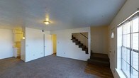 This two-story space is designed with practicality in mind. The main level features a carpeted living area with neutral white walls that lend a clean, understated aesthetic. A flush-mount ceiling light provides overhead illumination, while a doorway on one side leads to an adjacent room with warm yellow-toned walls, adding a subtle contrast to the otherwise neutral palette. The transition between carpet and hardwood flooring near the base of the staircase creates a natural visual boundary between areas. The staircase, positioned centrally within the space, features dark-toned carpeted steps with a simple metal railing, providing straightforward access to the upper floor. The textured ceiling adds a modest architectural detail throughout. The overall ambiance is functional and move-in ready, offering a blank canvas for personalization. The open arrangement of the main level allows for flexible furniture placement, making the space adaptable to a variety of living configurations.