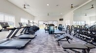Located in Hendersonville, NC, this fitness center offers a comprehensive exercise facility designed to support a wide range of workout routines. The single-floor space is thoughtfully laid out, with dedicated zones for cardio and strength training that allow for comfortable use without overcrowding.

The cardio section features a row of TRUE treadmills, stair climbers, and a rowing machine, all positioned to take advantage of the abundant natural light streaming through large windows and a full-length sliding glass door. The glass door opens to views of a landscaped outdoor area framed by a black iron fence, creating an airy, open atmosphere during workouts.

The strength training area is equally well-appointed, with a cable machine, adjustable benches, free weights with a dedicated rack, and additional resistance equipment. Full-length mirrors along one wall enhance the sense of space while supporting proper form during exercises.

Underfoot, modular carpet tiles add both comfort and a modern aesthetic. The vaulted ceiling, accented by ceiling fans and recessed lighting, contributes to the spacious and well-ventilated feel of the room. A water fountain station with dual dispensers is conveniently located near the entrance, and a wall-mounted TV provides entertainment during workouts. The overall design is clean, bright, and functional, making this a well-rounded fitness facility.