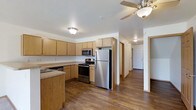 This two-floor space is designed with functionality and comfort in mind. The kitchen serves as a central feature, equipped with a full suite of stainless steel appliances, including a refrigerator, electric range, and built-in microwave. Light maple-toned cabinetry provides generous storage, complemented by a countertop with an integrated sink and a kitchen island that adds additional workspace and prep area. A ceiling fan overhead enhances air circulation and comfort in the space.

The flooring throughout is a warm hardwood-style material that adds continuity and a welcoming feel as it extends from the kitchen into the adjoining hallway and beyond. The hallway leads to additional rooms, offering a practical and open flow between spaces. Flush-mount ceiling lighting ensures consistent illumination throughout.

The overall layout is open and efficiently organized, with clean white walls that keep the space feeling bright and neutral. The combination of modern stainless appliances, warm wood tones, and straightforward design gives this two-floor space a practical yet inviting character suited for everyday living.