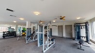 This fitness center is thoughtfully designed to accommodate a comprehensive range of workout activities. The space is divided into two distinct zones that flow seamlessly into one another. The cardio area is distinguished by a striking wood-paneled ceiling detail and an expansive bank of floor-to-ceiling windows that frame outdoor views and flood the space with natural light. A row of Matrix treadmills and elliptical machines lines this windowed wall, providing an energizing environment for cardio training.

The strength training area transitions naturally from the cardio zone and features a broad selection of cable machines, multi-functional weight stations, and benches suited for various resistance exercises. Full-length mirrors are strategically positioned throughout, enhancing the sense of openness while supporting proper form during workouts. Ceiling fans are distributed across both zones, contributing to air circulation and comfort during intense sessions.

The flooring throughout consists of durable, dark rubberized material appropriate for a high-use fitness environment. Recessed and flush-mount lighting supplements the natural light, ensuring consistent illumination across the entire space. Additional equipment including exercise balls and free weights can be seen stored along the perimeter, rounding out the facility's comprehensive fitness offerings.