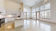 This contemporary single-floor space is defined by its striking architectural design and bright, open atmosphere. Polished concrete floors flow continuously throughout, lending a clean, modern aesthetic to every room. The entry corridor sets the tone immediately, with soaring ceilings and recessed wall niches creating a gallery-like passage into the main living area.

The open-concept living and kitchen space is a standout, anchored by dramatic floor-to-ceiling windows that wrap the corner of the room and offer expansive outdoor views. Two glass-paned doors provide direct access to a balcony, allowing natural light to pour in throughout the day. A ceiling fan adds comfort to this airy, voluminous space.

The kitchen is well-appointed with white shaker-style cabinetry, granite countertops, a subway tile backsplash with under-cabinet lighting, stainless steel appliances including a dishwasher and refrigerator, and a central island with pendant lighting overhead — ideal for both cooking and casual dining.

Additional rooms include a bright bedroom with tall windows and polished concrete floors, and a full bathroom featuring a tiled tub-shower combination, granite vanity, and white cabinetry. Throughout the space, the consistent use of neutral tones, high ceilings, and abundant natural light creates a cohesive, modern, and inviting environment.