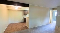 This single-story space offers a straightforward, functional layout that connects the living area to the kitchen through an open pass-through design. The living area is finished with neutral-toned carpet, providing a comfortable and understated foundation for furnishings. A partial dividing wall separates the two main zones while maintaining an open, airy feel between them.

The kitchen area features wood-look hard flooring, providing a practical and easy-to-maintain surface. Dark wood cabinetry lines the walls, offering ample storage space, and a countertop peninsula serves as a natural transition between the kitchen and living spaces. A ceiling fan is installed in the kitchen area, contributing to airflow and comfort throughout the unit.

The walls throughout are finished in a soft, neutral tone, complementing both the dark cabinetry and the light flooring materials. A textured ceiling adds subtle visual interest overhead. The overall design is classic and utilitarian, with an emphasis on functionality and ease of use. The open connection between living and kitchen areas makes the space feel cohesive and well-suited for everyday living.