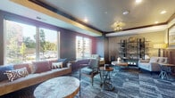 This thoughtfully designed single-floor amenity space is divided into three distinct areas, each offering its own character and function. The main lounge is a welcoming, light-filled gathering area featuring large windows that invite ample natural light and views of the surrounding neighborhood. Bold, deep-toned walls in charcoal and mauve are offset by gold-accented ceiling beams, creating a modern yet warm atmosphere. The space is furnished with a mix of mid-century modern and contemporary seating, including plush sofas, accent chairs, and stylish coffee tables atop patterned blue-toned carpeting. Decorative open shelving displays curated accent pieces, adding a gallery-like quality to the room. Adjacent to the lounge is a private conference or meeting room, distinguished by rich hardwood flooring, floor-to-ceiling dark wood wainscoting, and a striking wood-paneled ceiling with an ornate chandelier. A live-edge wooden conference table surrounded by soft velvet chairs anchors the room, while framed artwork and exposed white stone walls lend an upscale, intimate feel. The third area is a refined wine lounge, featuring built-in dark wood cabinetry with an integrated wine rack, leather club chairs, herringbone hardwood flooring, and a dramatic whitewashed stone accent wall — evoking an air of sophistication and relaxation.