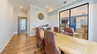 Located at 8651 Airway Dr in Pensacola, Florida, this single-room office is a thoughtfully designed professional workspace that balances functionality with stylish interior details. The space is finished with warm hardwood-style flooring throughout, crisp white walls, and crown molding that lends a polished, refined character to the room.

At the heart of the office sits a substantial rectangular conference table surrounded by cushioned brown leather executive chairs, making it well-suited for team meetings or client consultations. A wall-mounted flat-screen display is positioned nearby for presentations, while a dedicated workstation along one wall houses a computer and printer, supporting day-to-day operational needs. A large decorative Roman numeral clock serves as a striking focal point on the wall.

Natural light flows in through a large window dressed with bold patterned drapes and sheer white curtains, offering views of the surrounding landscape. A sleek black-framed glass sliding door connects the office to an adjacent interior space, maintaining visual openness while defining the room's boundaries. Recessed lighting and visible HVAC vents ensure a comfortable working environment year-round. The entrance is marked by a glass-panel exterior door with a clearly posted exit sign, meeting practical safety requirements. Overall, the space presents a professional yet inviting atmosphere ideal for collaborative work.