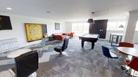 This single-floor recreational lounge is thoughtfully designed to serve as a dynamic social and leisure space. The room features a patterned carpet in shades of grey and blue, lending a modern, energetic feel to the interior. At the center of the space sits a full-size pool table with a striking pendant light overhead, making it a natural focal point for gatherings. A comfortable seating area with red sofas and accent chairs is arranged near large windows that invite an abundance of natural light and offer views of the surrounding greenery. Eclectic swivel chairs in black and blue are positioned throughout the room, providing flexible seating options for various activities. A large decorative wall piece — resembling an oversized Scrabble board — adds a playful, artistic touch to one wall, reinforcing the lounge's game-centric identity. The walls are crisp white, contrasted by a bold dark accent wall near the pool table area, creating visual depth. The space flows openly and generously, accommodating multiple activity zones without feeling cluttered. Overall, the lounge strikes a balance between casual comfort and contemporary design, making it a welcoming environment for recreation and social interaction.
