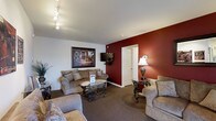 This single-floor space offers a warm and inviting atmosphere through thoughtful design and classic furnishings. The main sitting area serves as the focal point of the space, anchored by a rich, deep-red accent wall that adds a bold visual contrast to the otherwise neutral color palette of white walls and soft, carpeted flooring. Track lighting mounted on the ceiling provides flexible, adjustable illumination throughout the room, complementing the warm glow cast by a traditional table lamp in the corner. Seating is generous and comfortable, with a plush sofa and coordinating armchairs upholstered in a muted, textured fabric arranged around an ornate glass-top coffee table with decorative wrought iron legs. A matching side table with an elegant lamp adds symmetry to the layout. A flat-screen television is mounted on the accent wall, making the space as functional as it is stylish. A framed landscape painting adds a touch of artistic character to the wall. A doorway set into the accent wall leads to an adjoining room, hinting at additional functional space beyond. Overall, the space strikes a balance between classic elegance and everyday comfort.