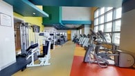 This fitness center offers a comprehensive workout environment designed to accommodate a wide range of exercise needs. The space is defined by its bold, energetic design — teal, yellow, and green accent panels line the walls and ceiling, creating a visually dynamic atmosphere that complements the active nature of the facility.

The layout is thoughtfully organized into distinct workout zones. A dedicated cardio area lines the windowed wall, featuring a row of treadmills, elliptical trainers, and stationary bikes. Tall, multi-pane windows flood this zone with natural light and offer views to the outdoors, making workouts feel open and inviting. Ceiling fans are mounted overhead to maintain airflow throughout the space.

The strength training section is equally well-appointed, offering a variety of cable machines, weight benches, leg press and extension equipment, and a fully stocked dumbbell rack displayed in front of floor-to-ceiling mirrors. The mirrors amplify the sense of space and allow users to monitor their form during exercises.

Underfoot, a multi-colored rubber flooring system — featuring sections of yellow, red, and gray — delineates different workout zones while providing comfort and safety. Recessed lighting throughout the teal-painted ceiling ensures consistent, even illumination across all areas. The overall ambiance is energetic, modern, and functional.