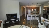 This single-floor space showcases a contemporary open-concept design that integrates the living and kitchen areas into one cohesive environment. The highly polished concrete floors extend throughout, reflecting light and contributing to a sleek, urban aesthetic.

The kitchen is a standout feature, equipped with dark espresso cabinetry, granite countertops, and stainless steel appliances including a full-sized refrigerator and built-in microwave. A large kitchen island with bar-height seating provides both additional prep space and a casual dining option. Above the island, three elegant pendant lights hang at varying heights, complementing the track lighting installed along the ceiling to ensure the kitchen is well-lit and functional.

The living area flows naturally from the kitchen, anchored by a flat-screen television on a low-profile media console. A plush area rug adds warmth and texture, contrasting nicely with the industrial quality of the concrete floors. A hallway leading to additional areas of the unit is visible, lined with framed artwork. The overall ambiance is modern, urban, and thoughtfully designed for open living.