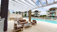 This well-appointed residential community offers an impressive array of outdoor amenities designed for comfort and leisure. At the heart of the property is a large, sparkling swimming pool surrounded by an expansive pool deck, where residents can relax on modern chaise lounges beneath open skies. A generous pergola-covered lounge area provides shade and shelter, furnished with contemporary outdoor seating that includes sofas, armchairs, and coffee tables — ideal for socializing or unwinding. An outdoor wet bar or kitchenette station is conveniently positioned nearby for added convenience. Adjacent to the pool deck, a glass-enclosed fitness center is equipped with cardio machines, including treadmills, and offers direct views of the pool area, allowing residents to exercise while enjoying the outdoor scenery. The complex is enclosed by clean white fencing for safety and privacy, with safety equipment such as a life ring readily accessible. Lush landscaping, including tropical palms and flowering plants, lines the covered walkways that connect the buildings, enhancing the property's resort-like ambiance. The multi-story residential buildings feature balconies and large windows, contributing to an open, airy aesthetic throughout the community.