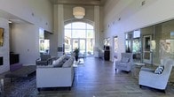 This impressive single-floor space makes an immediate impact with its dramatic, soaring ceilings that create a grand sense of openness and volume. The focal point of the main living area is a stunning floor-to-ceiling arched window arrangement that floods the interior with abundant natural light and offers unobstructed views of the lush outdoor surroundings, including mature palm trees and what appears to be a resort-style pool area beyond.

The interior design is clean and contemporary, featuring large-format tile flooring in a warm, neutral tone that complements the crisp white walls. Furnishings are tastefully selected, with a generous sectional sofa and coordinating accent chairs arranged to encourage conversation and relaxation. Decorative throw pillows and potted greenery add subtle texture and life to the space.

Flanking the central window wall, mirrored panels and glass sliding doors extend the visual depth of the room while enhancing the flow of natural light throughout. A large pendant light fixture anchors the ceiling above the seating area, providing both functional illumination and a refined design statement. The overall ambiance is bright, airy, and welcoming — a polished communal space designed for comfort and modern living.