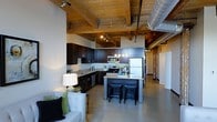 This single-floor urban loft blends industrial character with modern finishes to create a distinctive and functional living space. The open-plan layout seamlessly connects the kitchen and living area, fostering a sense of openness and flow throughout. Polished concrete floors extend across the entire space, providing a sleek, low-maintenance foundation that complements the industrial aesthetic.

The kitchen is a standout feature, boasting dark espresso cabinetry, light-toned stone countertops, and a full suite of stainless steel appliances, including a gas range, built-in microwave, and French door refrigerator. A central island with bar seating offers additional prep space and a casual dining option. Pendant lighting above the island adds warmth and focus to the work area.

Overhead, exposed wooden beams and original timber ceilings lend rustic warmth, while large-diameter industrial HVAC ductwork reinforces the loft's raw, urban character. Track lighting throughout the space provides flexible, directed illumination. The living area comfortably accommodates lounge seating and benefits from the open sightlines the layout affords. The overall ambiance is modern yet warm, striking a balance between raw industrial elements and refined contemporary finishes.