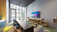 Located at 8446 S Dixie Hwy in Miami, Florida, this contemporary single-floor amenity lounge offers a well-designed, multi-functional communal space ideal for both relaxation and productivity. The room is anchored by soaring ceilings and a dramatic floor-to-ceiling glass curtain wall that floods the interior with natural light and frames views of the surrounding urban landscape.

The space is thoughtfully divided into distinct zones. A long, dark communal work surface lined with teal and neutral accent chairs runs along the window wall, providing an ideal setting for focused work or casual gatherings. Opposite, a large wall-mounted flat-screen TV is flanked by a series of floating natural wood open shelving units, adding warmth and a sense of organized display to the neutral gray walls.

A lounge seating area features a generous light gray sectional sofa adorned with colorful accent pillows, positioned around a sleek round side table — creating an inviting space for conversation or leisure. The floors are finished with a vibrant, geometric-patterned carpet in shades of green, yellow, gray, and brown, adding energy and visual interest throughout. Subtle pendant lighting complements the natural light, while touches of greenery, including a vertical plant wall accent near the entrance, bring a refreshing, biophilic element to the overall design.