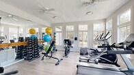 This fitness center offers a thoughtfully designed workout space that balances functionality with an open, airy atmosphere. Light-toned hardwood-style flooring runs throughout, complementing the neutral white and gray wall palette that gives the room a clean, modern feel. Large, multi-pane windows line the walls, flooding the interior with natural light and providing views of the surrounding landscaped outdoor area and pool beyond. Dual ceiling fans help maintain comfortable airflow throughout the space.

The room is comprehensively equipped to support a wide range of fitness activities. Cardio equipment includes treadmills, upright and recumbent stationary bikes, an elliptical, and a rowing machine. A dedicated free weights area features a full dumbbell rack alongside a weight bench, resistance bands, medicine balls, and stability balls in an array of sizes. A cable machine and wall-mounted TV add further versatility and entertainment. Floor-to-ceiling mirrors along one wall visually expand the space and assist with form during workouts.

A glass-paned door provides direct access to the outdoor pool area, seamlessly connecting indoor and outdoor amenities. French doors on an adjacent wall open to a neighboring lounge area, enhancing the overall flow and accessibility of the facility.