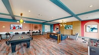 Located in Gonzales, Louisiana, this energetic game room delivers a bold, contemporary aesthetic across a generously sized single-floor layout. The interior is immediately distinguished by its striking color palette — salmon-pink and teal accent walls are complemented by teal-painted exposed ceiling beams, creating a lively and cohesive visual identity throughout the space.

The flooring features a vivid multicolored carpet with warm rust, gray, and blue tones that anchors the room's playful character. A full-size pool table serves as a central recreational focal point, while a foosball table provides additional entertainment options. Comfortable lounge seating, including upholstered armchairs and a sofa, is arranged around a flat-screen TV mounted on a warm wood-paneled wall, creating a relaxed viewing area.

A long, wall-mounted white counter with modern bar-height stools offers a dedicated workspace or casual bar seating area, complete with a mounted TV above. High-top bistro tables and stools are positioned near a sliding glass door that fills the corner with natural light and provides outdoor views of the surrounding area. A sculptural multi-arm chandelier and gold-toned sconce lighting add refined decorative touches. A distinctive arched niche on a bold red accent wall frames a piece of abstract wall art, further elevating the room's design-forward atmosphere.