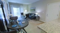 This single-floor residence showcases an open-concept layout that seamlessly connects the dining, living, and kitchen areas into one cohesive space. The dining area is anchored by a round dark-wood table with seating, positioned adjacent to a kitchen counter finished in granite, offering a natural transition between meal preparation and dining. The living area is generously proportioned, featuring plush wall-to-wall carpeting that adds warmth underfoot and a neutral color palette throughout. A blue sofa serves as the centerpiece of the living space, complemented by accent chairs and dark-finished side tables, contributing to a cohesive and inviting atmosphere. Sliding glass doors along the far wall flood the interior with natural light and provide direct access to a private balcony overlooking lush outdoor greenery. Decorative curtains frame the doors, adding a refined touch. The walls are finished in a soft, neutral tone, enhancing the brightness of the space. Recessed lighting and floor lamps throughout provide layered illumination, making the overall ambiance both functional and welcoming.