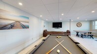 This single-floor clubhouse is designed to serve as a central gathering and recreational hub for its community. The open layout features light vinyl plank flooring throughout, a drop tile ceiling with recessed lighting, and a clean, neutral color palette of soft blues and greens that lend a relaxed, coastal atmosphere — complemented by nautical-themed wall art.

The game area is a highlight of the space, anchored by a full-size pool table and a shuffleboard lane running along the wall, offering ample entertainment options. Adjacent to the game area, a comfortable lounge zone features rich brown leather sofas and armchairs arranged around a large wall-mounted flat-screen television with a soundbar, creating an inviting setting for watching sports or socializing.

A rustic high-top wooden table with bar stools is positioned near a row of large windows and a set of glass double doors, which allow natural light to fill the space and provide views of the surrounding greenery outside. This area also serves as a casual spot for seating or informal gatherings.

Additional functional amenities are tucked toward the rear of the space, including an in-unit washer and dryer, accessible through a dedicated utility area. Picnic-style tables are also visible, adding flexible seating for group use. The overall design balances practicality with comfort, making this clubhouse a versatile and welcoming community amenity.