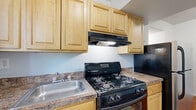 This single-floor space offers a practical and well-organized layout across its key living areas. The kitchen is a standout feature, outfitted with light natural wood cabinetry that provides generous storage above and below the counter. Granite-look countertops complement the cabinetry, while a black gas range with an overhead exhaust hood serves as the cooking centerpiece. A stainless steel refrigerator and a single-basin stainless steel sink complete the kitchen's functionality. A window overlooks greenery, bringing in natural light and a connection to the outdoors. The kitchen also houses a water heater, and durable tile-style flooring runs throughout the space.

The bedroom showcases warm, polished parquet wood flooring in a classic geometric pattern that adds character and visual interest to the room. Crisp white walls and a window allowing natural light give the room an open, airy feel. A flush-mount ceiling fixture provides overhead lighting.

The bathroom features a full-size soaking tub with a tile surround in clean white, complemented by a window with blinds that allows for natural ventilation and light. The overall aesthetic of the space is clean and straightforward, offering a comfortable and functional environment.