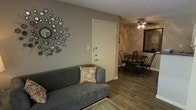This single-story space presents an open-concept layout that seamlessly connects the living and dining areas, creating a cohesive and functional flow. The living area features a plush gray sofa accented with a decorative patterned pillow, complemented by a starburst mirror wall accent that adds a stylish focal point to the neutral-toned walls. The wood-look flooring extends throughout the visible space, providing warmth and visual continuity between rooms.

The dining area, visible from the living room, accommodates a round table surrounded by classic Windsor-style black chairs, offering an intimate setting for meals. A ceiling fan with an integrated light fixture serves the dining space, providing both air circulation and ambient lighting. The textured ceiling adds a traditional architectural detail consistent throughout the space.

The entryway, positioned between the two areas, features a clean white door with standard hardware, creating a clear transition point. Neutral beige and gray wall tones are used throughout, contributing to a calm and cohesive atmosphere. A large framed mirror in the dining area enhances the sense of openness. Overall, the space balances practicality with understated decorative touches.