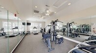 Located at 859 S Cliffs Cir in Spring Lake, NC, this fitness center offers a dedicated exercise room designed to support a comprehensive workout routine. The space is finished with neutral-toned walls, carpeted flooring, and a tray ceiling accented by recessed lighting and dual ceiling fans, creating a bright and comfortable environment for exercise.

The room is outfitted with a diverse selection of equipment to accommodate both cardio and strength training. Cardio options include a treadmill, elliptical trainer, and stationary bike, while strength training is supported by a multi-station cable machine, a weight bench, and a full rack of dumbbells ranging across multiple weight increments. A wall-length panel of mirrors runs along one side of the room, visually expanding the space and allowing users to monitor their form during workouts.

Natural light filters in through multiple windows fitted with white shutters, contributing to an open and airy atmosphere. A wall-mounted television provides entertainment during workouts, and a bulletin board near the entry door offers a space for community notices. A dedicated entrance door with an exterior view adds convenient access. The overall design balances functionality with a clean, well-maintained aesthetic suited for regular fitness use.