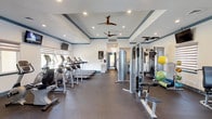 This fitness center is designed to support a comprehensive workout experience within a spacious, single-floor layout. The main exercise area features an impressive lineup of cardio equipment, including multiple treadmills and elliptical machines, all neatly arranged to maximize floor space and ease of movement. Wall-mounted televisions are positioned for viewing during cardio sessions, adding to the overall comfort of the space.

Strength training is well-represented, with a multi-station cable machine and a dedicated squat rack with an adjustable bench, catering to a full range of resistance exercises. A selection of stability balls and resistance accessories are also available, supporting functional and flexibility training. The flooring throughout is a durable rubber material, providing both safety and comfort underfoot.

Architecturally, the space is defined by elegant tray ceilings with blue accent molding and recessed lighting, creating a polished and inviting atmosphere. Dual ceiling fans promote air circulation throughout. Natural light filters in through zebra-shaded windows, complementing the bright white walls. A set of glass double doors at the entrance offers views to the exterior and contributes to the open, airy feel. The overall design balances functionality with a refined aesthetic, making this fitness center both practical and welcoming.