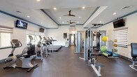 This fitness center is designed to support a comprehensive workout experience within a spacious, single-floor layout. The main exercise area features an impressive lineup of cardio equipment, including multiple treadmills and elliptical machines, all neatly arranged to maximize floor space and ease of movement. Wall-mounted televisions are positioned for viewing during cardio sessions, adding to the overall comfort of the space.

Strength training is well-represented, with a multi-station cable machine and a dedicated squat rack with an adjustable bench, catering to a full range of resistance exercises. A selection of stability balls and resistance accessories are also available, supporting functional and flexibility training. The flooring throughout is a durable rubber material, providing both safety and comfort underfoot.

Architecturally, the space is defined by elegant tray ceilings with blue accent molding and recessed lighting, creating a polished and inviting atmosphere. Dual ceiling fans promote air circulation throughout. Natural light filters in through zebra-shaded windows, complementing the bright white walls. A set of glass double doors at the entrance offers views to the exterior and contributes to the open, airy feel. The overall design balances functionality with a refined aesthetic, making this fitness center both practical and welcoming.