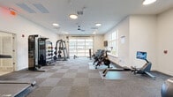 Located at 2755 Red Bloom Dr in Greenwood, Indiana, this fitness facility is thoughtfully designed across two exercise rooms, offering a comprehensive workout environment for residents. The main exercise room is bright and open, featuring a large glass garage door that floods the space with natural light and provides views of the surrounding community. Checkered carpet tile flooring covers the area, which is outfitted with an extensive array of equipment including spin bikes, a rowing machine, a cable machine, a multi-functional strength rack, medicine balls, and a full dumbbell rack ranging across multiple weight increments. A wall-mounted television and ceiling fan add comfort and convenience. The secondary exercise room is separated by sleek floor-to-ceiling glass partition walls with black metal framing, creating a clean, modern aesthetic. This studio space houses three treadmills with interactive touchscreen displays, a functional training rack with TRX suspension straps and kettlebells, a stability ball, and a wall mirror. Recessed lighting and ceiling fans are present throughout both rooms, maintaining a well-lit and comfortable atmosphere. Together, the two rooms provide a versatile fitness experience suited for cardio, strength training, and functional movement workouts.