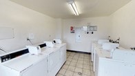 This single-floor shared laundry facility is designed with practicality and efficiency in mind. The room features light-colored walls and neutral tile flooring that contribute to a bright, clean atmosphere. Overhead fluorescent lighting ensures the space is well-lit throughout. Multiple top-loading and front-loading washing machines are arranged along the walls, complemented by a matching set of dryers, providing ample capacity for users. An electrical panel is mounted on the far wall, and informational notices and posted instructions are displayed nearby for user guidance. The layout is open and easy to navigate, allowing multiple users to access machines simultaneously without crowding. The overall design prioritizes function and cleanliness, making it a reliable and accessible laundry resource.