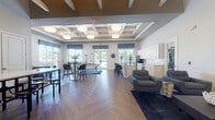 This well-appointed single-floor clubhouse is designed to serve as a dynamic social hub, offering a variety of activity and relaxation zones within one expansive, open-concept layout. The main gathering area is anchored by a striking coffered ceiling with recessed lighting and large pendant fixtures, lending architectural character and an elevated sense of height to the space. Warm herringbone-patterned hardwood flooring flows seamlessly throughout, tying each zone together cohesively.

The central open area accommodates a full-sized pool table and multiple high-top dining tables with bar seating, making it ideal for casual entertainment and socializing. A dedicated lounge area features coordinated blue patterned upholstered chairs and sofas arranged around a flat-screen television, creating a comfortable space for gathering and viewing. Decorative wall art and sculptural accents contribute to the polished, modern aesthetic carried throughout.

A game and activity room includes a long communal table with fixed seating and wall-mounted games, providing a dedicated space for recreational use. An additional seating nook with a card or game table offers further flexibility for group activities.

The full catering-style kitchen is outfitted with white shaker cabinetry, quartz countertops, a subway tile backsplash, stainless steel appliances, and a window overlooking the outdoor pool area. Large windows throughout the clubhouse invite abundant natural light and outdoor views, enhancing the overall bright and welcoming ambiance.