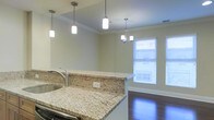 This single-floor space showcases an open-concept layout that seamlessly connects the kitchen and living areas, creating a cohesive and functional environment. The kitchen is highlighted by elegant granite countertops with distinctive veining in warm tones of beige and gold, complemented by a stainless steel sink and modern fixtures. A raised breakfast bar serves as a natural divider between the kitchen and the adjoining living area, offering a casual dining or entertaining option.

Overhead, a series of pendant lights hang above the kitchen workspace, providing focused task lighting while adding a contemporary design element. Crown molding lines the ceiling throughout, lending a refined architectural detail to the space. The transition into the living area reveals rich, dark hardwood flooring that adds warmth and contrast to the neutral wall tones.

A window fitted with white horizontal blinds allows natural light to filter into the living area, brightening the space. A small chandelier above the dining or living zone adds an additional layer of ambient lighting. The overall aesthetic is clean, modern, and well-appointed, with consistent finishes throughout that create a polished and inviting atmosphere.