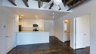 This single-story space showcases a thoughtfully designed open-concept layout that seamlessly connects the kitchen and living areas. The kitchen is outfitted with white cabinetry, a subway tile backsplash, stainless steel appliances including a microwave and range, and a breakfast bar with a light wood countertop that provides both workspace and casual seating.

One of the most striking architectural features is the exposed beam ceiling, where dark rustic wood beams contrast beautifully against the bright white ceiling, adding character and visual depth to the space. A ceiling fan is centrally positioned, enhancing comfort and airflow. Polished dark concrete floors run throughout, offering a durable, industrial-chic aesthetic that grounds the otherwise light and airy interior.

The open hallway leads to additional rooms, including what appears to be a bathroom with vanity cabinetry visible in the background, as well as closet storage. Recessed lighting and pendant fixtures provide layered illumination throughout. The crisp white walls and clean lines create a minimalist, contemporary ambiance, making the space feel open, functional, and move-in ready.