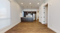 This single-floor space showcases a cohesive modern design defined by clean lines, white walls, and warm wood-tone flooring that runs throughout the main living areas. The open-concept layout connects the living area to the kitchen seamlessly, creating an expansive, airy feel enhanced by recessed ceiling lighting.

The kitchen is a standout feature, equipped with dark espresso flat-front cabinetry extending to the ceiling, a white quartz peninsula with an undermount sink, a textured tile backsplash, and stainless steel appliances including a French door refrigerator and built-in microwave. A trio of modern pendant lights hangs above the peninsula, adding both function and style.

The living area benefits from large windows fitted with roller shades, allowing ample natural light while maintaining privacy. The bedroom is carpeted and bright, with a window and recessed lighting providing a comfortable retreat.

The bathroom continues the contemporary aesthetic with a floating dark wood vanity, a wide framed mirror, matte black hardware and fixtures, and wood-look flooring. The entryway features white panel doors with matte black hardware, completing the space's modern, unified look.