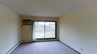 This single-floor space features a straightforward, functional layout centered around a carpeted living area. The room is finished with light-colored walls and a smooth ceiling, creating a neutral and airy atmosphere. A prominent sliding glass door serves as the focal point of the main living area, allowing natural light to fill the space and providing direct access to a balcony that overlooks a green outdoor area with trees and open grounds. Sheer curtains frame the sliding door, offering privacy while maintaining the flow of natural light. A wall-mounted air conditioning unit is positioned adjacent to the sliding door, ensuring climate control throughout warmer months. Baseboard heating runs along the perimeter of the room, providing an efficient heating solution during cooler seasons. The carpeted flooring adds a layer of comfort and warmth underfoot. The overall ambiance is clean and simple, with minimal decorative elements that allow the space to serve as a blank canvas for personalization. The connection to the outdoor balcony and the natural views beyond are standout features of this space.