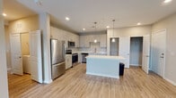 This well-designed single-floor residence offers a bright, contemporary living experience anchored by an open-concept layout. The kitchen is a standout feature, showcasing gray shaker-style cabinetry, a large central island with a quartz countertop, stainless steel appliances including a refrigerator, range, microwave, and dishwasher, and stylish pendant lighting overhead. A built-in pantry with open shelving adds practical storage. Light wood-look flooring flows throughout the main living areas, complementing the neutral, warm-toned walls and clean white trim. The living area is generously proportioned and benefits from natural light through multiple windows, with a ceiling fan overhead. The bedrooms are carpeted for comfort, each featuring ceiling fans and windows that invite natural light. One bedroom offers a particularly bright corner window arrangement with outdoor views. The two bathrooms are finished with wood-look tile flooring and large-format stone-tiled surrounds. The primary bathroom features a walk-in glass-enclosed shower, an extended vanity with quartz countertop, and a large mirror. The secondary bathroom includes a full soaking tub with a tiled surround and a white vanity. Throughout, the space maintains a cohesive, modern aesthetic with consistent finishes and thoughtful design details.