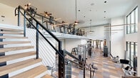 Located in Palm Harbor, Florida, this expansive two-story fitness facility delivers a premium workout experience through thoughtful design and comprehensive amenities. The main floor sets an impressive tone with soaring double-height ceilings, large floor-to-ceiling windows, and exposed industrial-style ductwork that adds an urban aesthetic. Patterned carpet tiles in warm neutral tones flow throughout the gym areas, while herringbone wood-look flooring defines the welcoming entry zone, accented by a lounge chair and sleek room dividers with dark-framed glass panels. The main training floor is equipped with a wide array of strength machines, cable systems, free weights, and functional training rigs. A dedicated functional fitness room features a power rack, punching bag, medicine balls, and an air bike, opening to the outdoors via a full glass garage-style door. A striking open-riser staircase with cable railings and wood treads connects both levels, serving as an architectural centerpiece. The upper mezzanine level hosts a dedicated cardio zone lined with stair climbers, ellipticals, and other aerobic equipment, all beneath ceiling fans and pendant lighting. Throughout both floors, curated wall art and teal accent walls contribute to an energizing yet polished atmosphere.
