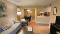 This single-floor space showcases an open-concept design that connects the living, dining, and kitchen areas into one cohesive and functional layout. The living area features warm hardwood flooring, neutral taupe-toned walls, and comfortable seating accented by a sleek arc floor lamp, creating a welcoming and relaxed atmosphere. A dark wood coffee table anchors the space, adding a grounded contrast to the light grey upholstered sofa and accent chair.

The living area flows naturally into the dining room, which is defined by a classic chandelier overhead and a dark wood dining table with matching chairs, providing an inviting spot for shared meals. The warm wall tones in this area add depth and a cozy, intimate feel.

The kitchen is accessible via a raised breakfast bar with granite-style countertops, offering both casual seating and a visual division from the main living space. Wood-toned cabinetry and stainless steel appliances lend a practical yet stylish finish. Throughout the space, consistent hardwood flooring ties each area together seamlessly, enhancing the sense of openness and continuity across the entire floor plan.