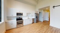 This single-floor unit showcases a contemporary design highlighted by notably tall ceilings and light wood-style flooring that flows consistently throughout the space. The kitchen is a standout feature, outfitted with crisp white shaker cabinetry extending to ceiling height, bright white countertops, and a full suite of stainless steel appliances including a French door refrigerator with water dispenser, electric range, built-in microwave, and dishwasher. A single-basin sink with a modern gooseneck faucet anchors the countertop workspace, and an open shelving nook provides additional storage. The living area benefits from dramatic floor-to-ceiling windows framed in dark metal, flooding the room with natural light and offering expansive views of the surrounding green landscape. The bedroom is generously proportioned with clean white walls and ample space for furnishings. The bathroom maintains the unit's clean aesthetic with white shaker cabinetry, a white countertop vanity with an illuminated mirror, and a full tub-shower combination finished in white tile. Throughout, bright white walls and a neutral palette create a cohesive, modern ambiance well-suited for comfortable everyday living.