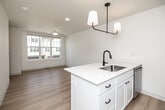 This thoughtfully designed three-story residence offers a clean, modern aesthetic throughout, with light hardwood flooring, crisp white walls, and black hardware accents tying each level together cohesively. The ground floor houses a utility room and a spacious attached garage with a concrete floor, automatic opener, and ample room for storage or parking. Staircases with warm wood handrails connect each level, creating a smooth flow through the home. The main living floor features an open-concept kitchen and living area bathed in natural light. The well-appointed kitchen is outfitted with white shaker cabinetry, quartz countertops, stainless steel appliances including a refrigerator, dishwasher, microwave, and range, plus a striking black pendant light fixture overhead. The top floor is dedicated to the private bedroom suite, which is generously sized and features a ceiling fan with integrated light, large windows with neighborhood views, and direct access to a walk-in closet with built-in shelving. The full bathroom is finished in white with clean-lined cabinetry, a white countertop vanity, and matte black and brushed gold accents. A hallway connects the upper-level rooms, completing this efficient and stylish multi-floor home.