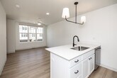 This thoughtfully designed three-story residence offers a clean, modern aesthetic throughout, with light hardwood flooring, crisp white walls, and black hardware accents tying each level together cohesively. The ground floor houses a utility room and a spacious attached garage with a concrete floor, automatic opener, and ample room for storage or parking. Staircases with warm wood handrails connect each level, creating a smooth flow through the home. The main living floor features an open-concept kitchen and living area bathed in natural light. The well-appointed kitchen is outfitted with white shaker cabinetry, quartz countertops, stainless steel appliances including a refrigerator, dishwasher, microwave, and range, plus a striking black pendant light fixture overhead. The top floor is dedicated to the private bedroom suite, which is generously sized and features a ceiling fan with integrated light, large windows with neighborhood views, and direct access to a walk-in closet with built-in shelving. The full bathroom is finished in white with clean-lined cabinetry, a white countertop vanity, and matte black and brushed gold accents. A hallway connects the upper-level rooms, completing this efficient and stylish multi-floor home.