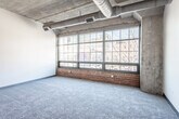 This single-floor unit showcases a compelling industrial aesthetic rooted in its original architectural character. The main living area is generously proportioned, featuring an exposed concrete ceiling with visible ductwork running overhead, raw concrete structural columns, and plush grey carpeting underfoot. A striking bank of steel-framed, factory-style windows spans a full wall, set atop a red brick base, flooding the space with natural light and offering views of the surrounding urban streetscape.

The open floor plan connects the living area to a compact, functional kitchen equipped with white cabinetry, dark laminate countertops, a range with oven, and wood-look vinyl flooring. A cylindrical concrete column serves as a distinctive architectural accent near the transition between spaces.

The bedroom is a well-sized, carpeted room with tall ceilings, offering a sense of openness reinforced by the unit's loft-inspired design. A closet provides practical storage, and a door connects to the hallway. The bathroom features wood-look vinyl flooring, a vanity with a yellow countertop and single sink, a toilet, and a bathtub with overhead grab bars. Throughout the unit, the interplay of raw industrial materials and clean white walls creates a bold, urban ambiance.