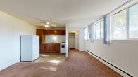 This single-floor studio offers an efficient, open-concept layout that combines the living and kitchen areas into one cohesive space. The living area is finished with wall-to-wall carpeting and features a ceiling fan overhead, a baseboard heating unit along the wall, and a window dressed with sheer curtains that allows natural light to filter into the room. The entry door opens directly into this main living space, which provides a straightforward, functional flow throughout.

The kitchen is defined by warm-toned wood cabinetry providing ample upper and lower storage, a light-colored countertop with an integrated sink, a white gas range, and a full-size white refrigerator. Tile flooring distinguishes the kitchen zone from the carpeted living area. A walk-in closet is conveniently situated just off the main living space, offering notable storage capacity for a studio layout.

The bathroom includes a full tub-and-shower combination with gray tile surround, a white vanity with under-cabinet storage, a large mirror, and tiled flooring. The overall design of the space is practical and straightforward, making efficient use of its footprint while providing all essential amenities within a single-level layout.