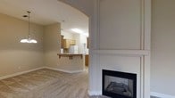 This single-floor residence offers a thoughtfully designed open layout that promotes a sense of ease and connectivity throughout the living spaces. The living area serves as the heart of the home, anchored by a classic fireplace framed with a crisp white mantel and paneled surround, providing both a focal point and a cozy gathering spot. Warm, neutral-toned carpet flows underfoot, complementing the soft beige walls that lend a calm, inviting ambiance to the space.

An elegant arched passageway connects the living area to the kitchen, adding an architectural detail that enhances the open feel while subtly defining each space. The kitchen is well-appointed with warm wood-toned cabinetry, a white refrigerator, and a raised breakfast bar with a dark wood countertop overhang — ideal for casual dining or entertaining. Tile flooring in the kitchen provides a practical contrast to the carpeted living area. Overhead lighting illuminates the kitchen efficiently, keeping the space bright and functional.

Overall, the residence presents a clean, neutral palette throughout, making it a versatile canvas adaptable to a variety of interior styles. The seamless flow between living and kitchen areas, combined with the warmth of the fireplace and the charm of the arched entryway, creates a welcoming and comfortable living environment.