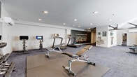 This fitness center offers a comprehensive workout environment designed to support a wide range of exercise routines. The space is thoughtfully organized into distinct functional zones, with plush, patterned carpet tiles covering the main floor area and durable rubber flooring underlining the weight and equipment sections. Natural light streams in through multiple windows along the perimeter walls, complementing the recessed ceiling lighting that keeps the room bright and energizing throughout.

The cardio zone features multiple treadmills, elliptical trainers, and spin bikes — each equipped with individual monitors for entertainment or performance tracking. A dedicated free weights area includes a full rack of dumbbells in graduated sizes, a multi-station cable machine, adjustable workout benches, and a stability ball, offering versatile strength training options. A large wall mirror expands the visual depth of the space and assists with form checks during exercise, while wall-mounted flat-screen TVs provide added entertainment.

A standout feature is the bold, large-scale mural on a deep green accent wall that reads "It's Nice to Have You in BHAM," giving the space a local character and welcoming energy. A water station is also conveniently available. The overall ambiance is modern and motivating, making this fitness center a fully functional and visually engaging place to work out.