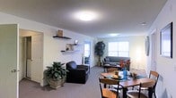 Located at 2230 Greenville Blvd in Greenville, NC, this single-floor space offers a clean, functional layout designed for comfortable everyday living. The open-concept main area seamlessly combines a dining zone and a living room, creating a spacious and connected feel throughout. The dining area accommodates a full-sized table and chairs, making it well-suited for meals and gatherings. The living area features plush carpeted flooring, comfortable seating, and wall-mounted shelving that adds both storage and a decorative touch. Multiple windows allow generous natural light to fill the space, enhancing the bright and airy atmosphere. Neutral wall tones provide a versatile backdrop throughout, complemented by tasteful greenery that adds warmth and life to the interior. The textured ceiling and flush-mount lighting fixtures contribute to the practical, no-fuss aesthetic of the space. A doorway visible in the image suggests additional rooms or storage beyond the main living area. Overall, this space presents a well-organized, inviting environment that balances functionality with comfort, making it a practical choice for those seeking a straightforward and livable layout in Greenville, NC.