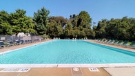 This outdoor pool facility centers around a generously sized swimming pool with crystal-clear turquoise water, a concrete surround deck, and clearly marked depth indicators beginning at a shallow three-foot entry point. The pool is framed on all sides by a wide, light-toned concrete deck that provides ample space for circulation and relaxation. Rows of lounge chairs and reclining chaise seating line the perimeter, offering poolside comfort beneath open skies. A lush backdrop of mature deciduous and evergreen trees surrounds the fenced enclosure, creating a natural, shaded boundary that enhances privacy and ambiance.

Adjacent to the pool area is a single-story support building constructed of painted concrete block. The structure includes utility access doors and a covered walkway along its exterior, where additional seating is available. The building's interior spaces are functional and utilitarian, with painted block walls and concrete flooring, housing what appears to be storage and mechanical or restroom facilities. The covered overhang along the building exterior provides a shaded transition zone between the support structure and the open pool deck, making it a practical and well-organized outdoor recreational space.