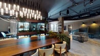 Located at 714 N Racine Ave in Chicago's vibrant West Town neighborhood, this thoughtfully designed single-floor amenity space masterfully blends industrial aesthetics with refined, contemporary interiors. Exposed ceiling infrastructure and dark overhead elements contrast beautifully with warm wood accents, polished concrete floors, and soft textile finishes throughout.

The space is organized into several distinct yet flowing zones. A striking communal dining area anchors one section, featuring a substantial solid-wood table surrounded by cream upholstered chairs and illuminated by a dramatic cascade of pendant lighting. Lush greenery, including large fern arrangements integrated into the furniture, adds a vibrant, organic energy to the space.

Adjacent to the dining area, a full-service kitchen bar with deep blue cabinetry and open display shelving offers a polished hospitality-style setup complete with screens for entertainment. A lounge zone transitions seamlessly nearby, offering generous light-gray sofas, a slatted wood ceiling detail, and a curved architectural partition that adds visual interest.

Tucked-away booth seating with arched alcoves and warm pendant lighting provides intimate gathering options, while private workspaces with floor-to-ceiling windows offer views of an outdoor terrace. The overall ambiance is sophisticated yet welcoming, balancing community-focused gathering with quieter, more focused areas.